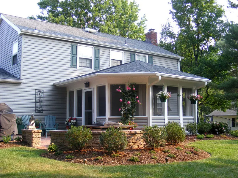 A grey clapboard house with a screened front porch, flowering baskets and red chairs on the lawn