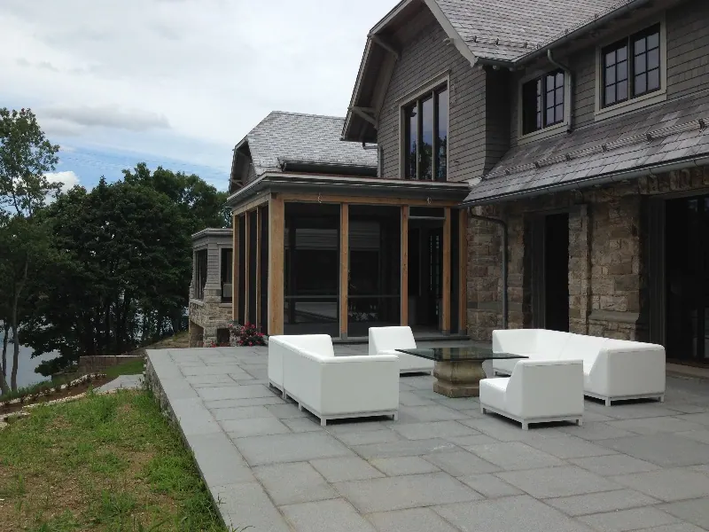 A shingled house with a screened porch opening onto a bluestone patio with white outdoor furniture