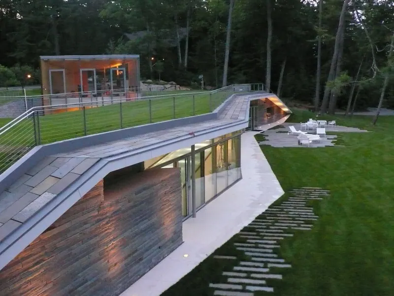 A modern house at dusk beside a fenced sports court, with a screened structure under a sloping roof