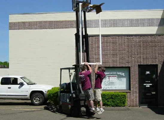 Two men lifting an oversized Screen Wall panel with a forklift outside the shop