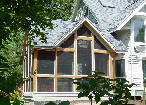 A shingled house with a timber-framed screened porch surrounded by foliage